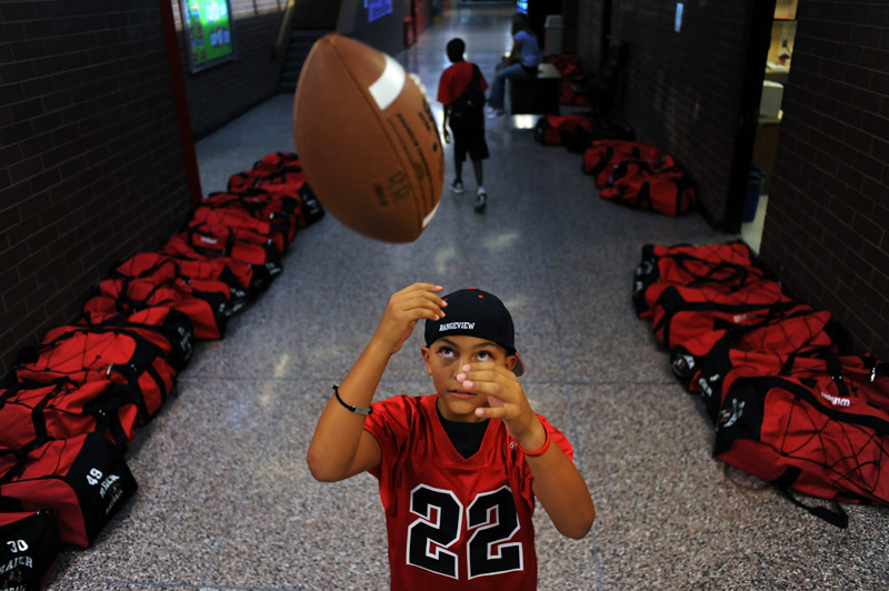 Rangeview High School ball boy and son of head coach Dave Gonzales, Max Gonzales, 10, plays with a football in the hallway as the players bags can be seen lining the walls before the team played Gateway High School  Friday 09/11/09 at Aurora Public School Stadium.  Photo by Matt McClain